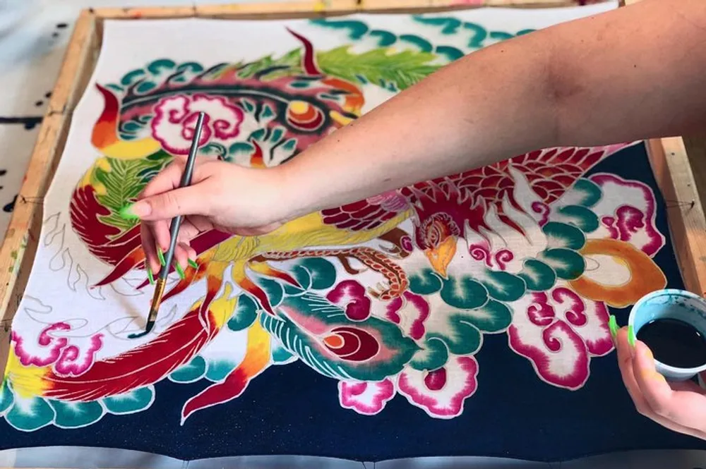 An artists hand is shown painting a vibrant and detailed phoenix on fabric stretched over a frame