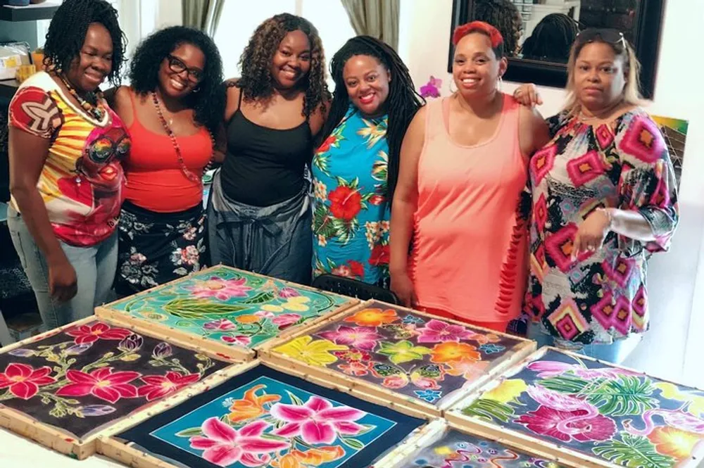A group of six smiling women are standing behind a table displaying vibrant floral paintings