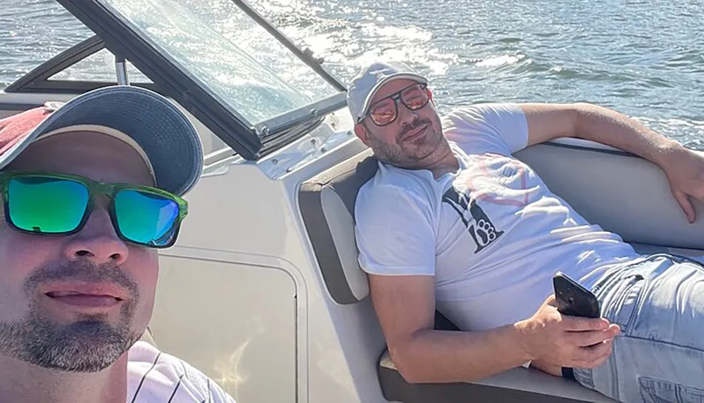 Two men are enjoying a sunny day on a boat with one taking a selfie and the other lounging in the background