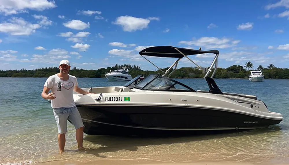 A man is standing on a beach next to a moored motorboat gesturing towards himself with a confident pose with tropical vegetation and boats in the background