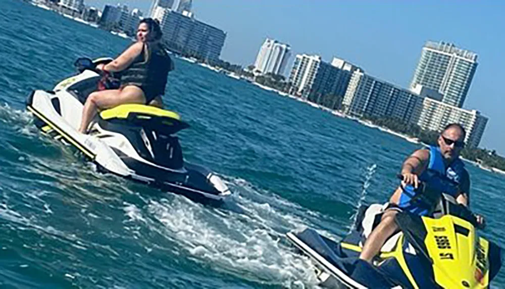 Two people are riding jet skis on the water with coastal buildings in the background