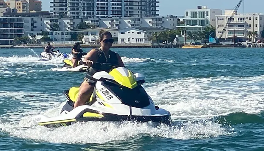 Several people are riding jet skis on a sunny day near a coastline with buildings in the background.