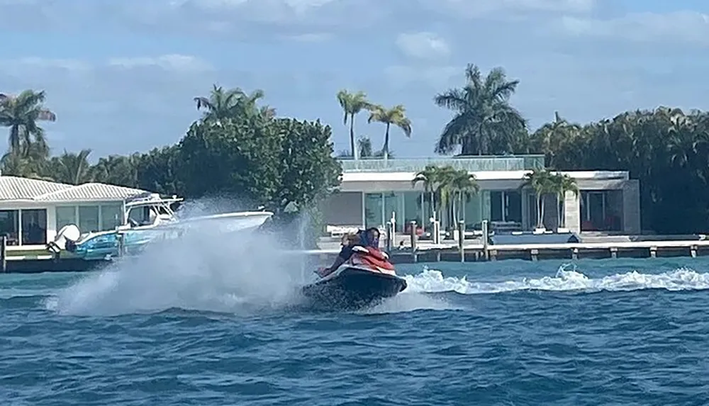 A person is riding a jet ski at high speed near what appears to be a waterfront property with palm trees and docks