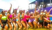 A group of people in swimwear is cheerfully posing in shallow water with drinks in their hands, wearing matching visors, and some of them are holding up a cutout of a person's face.