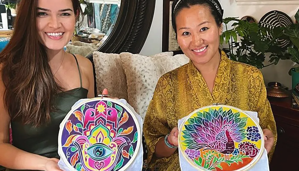 Two smiling women are showing off their colorful patterned artwork held in embroidery hoops