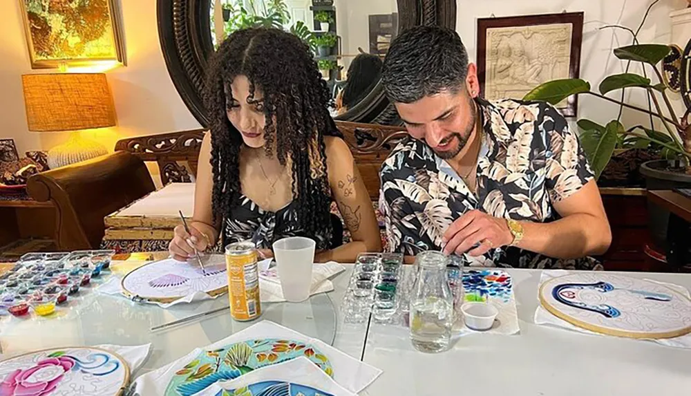 Two individuals are focused on painting colorful designs on embroidery hoops at a table covered with art supplies