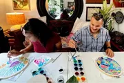 Two individuals are engaged in painting on oval-shaped canvases at a table adorned with painting supplies, exhibiting a creative and enjoyable activity.