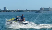 A person is riding a jet ski across a body of water with a city skyline in the background.