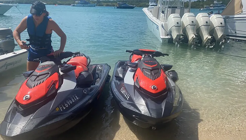 A person in a life vest stands beside two jet skis on the waters edge with a boat equipped with multiple outboard engines in the background