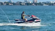 A person is riding a jet ski on a sunny day with a coastal city skyline in the background.