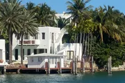 A white, modern-style villa is nestled among palm trees along the waterfront with a private dock in the foreground.