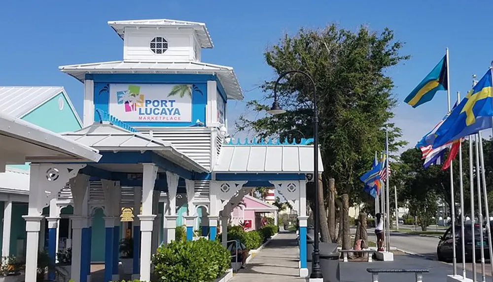 The image features a sunny view of the Port Lucaya Marketplace entrance with a row of vibrant international flags lining the street