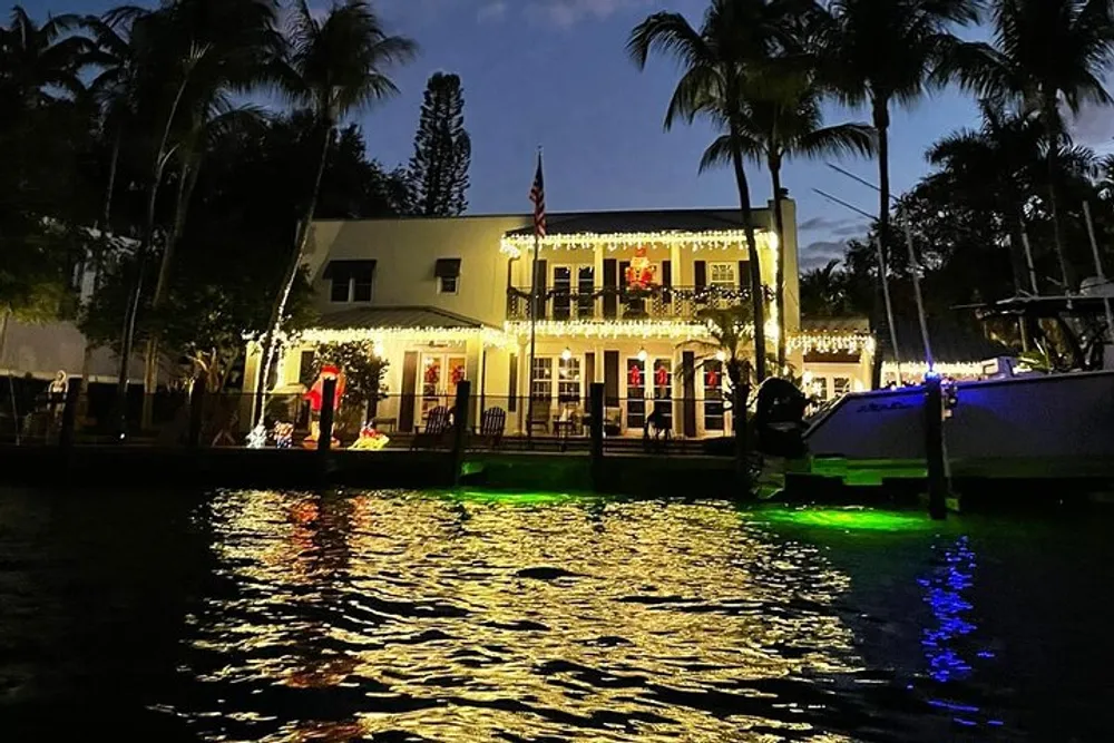 The image depicts a two-story house adorned with festive lights including a lit Christmas tree on the balcony situated beside a body of water reflecting the colorful lights at dusk