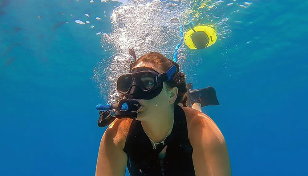 A person is snorkeling underwater exhaling bubbles near a floating yellow snorkel tube