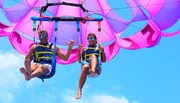 Two people are enjoying a parasailing adventure in a clear blue sky with a vibrant pink parachute above them.