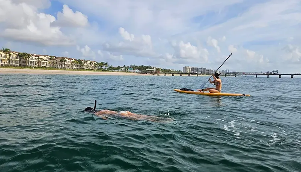 A person is paddleboarding near the shoreline while another person appears to be swimming or snorkeling in the sea nearby