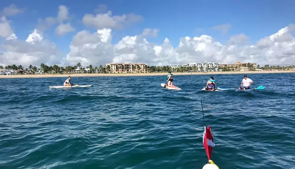 A group of people are paddleboarding on a calm blue sea near a coastal area with buildings in the background under a sky dotted with clouds