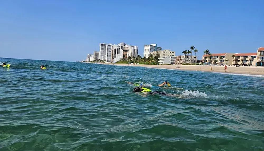 Swimmers wearing bright swim caps are seen in the ocean near a sandy beach lined with buildings on a sunny day