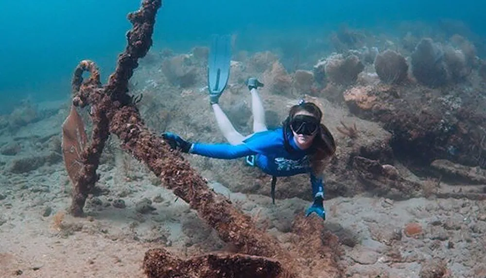 A person is free-diving underwater near a large corroded anchor surrounded by marine growth
