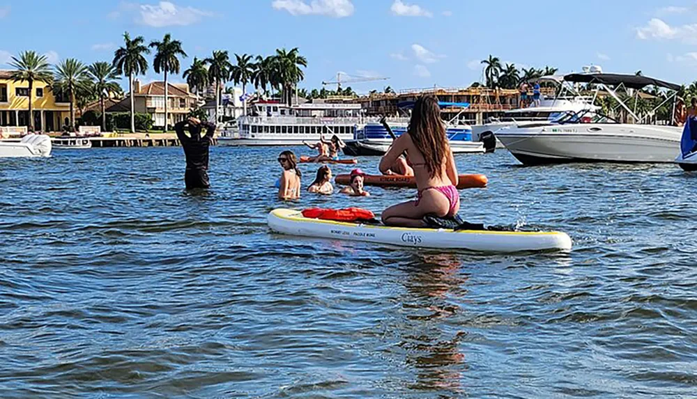 A group of people are enjoying water activities on a sunny day with some standing in the water a girl sitting on a paddleboard and luxurious houses in the background