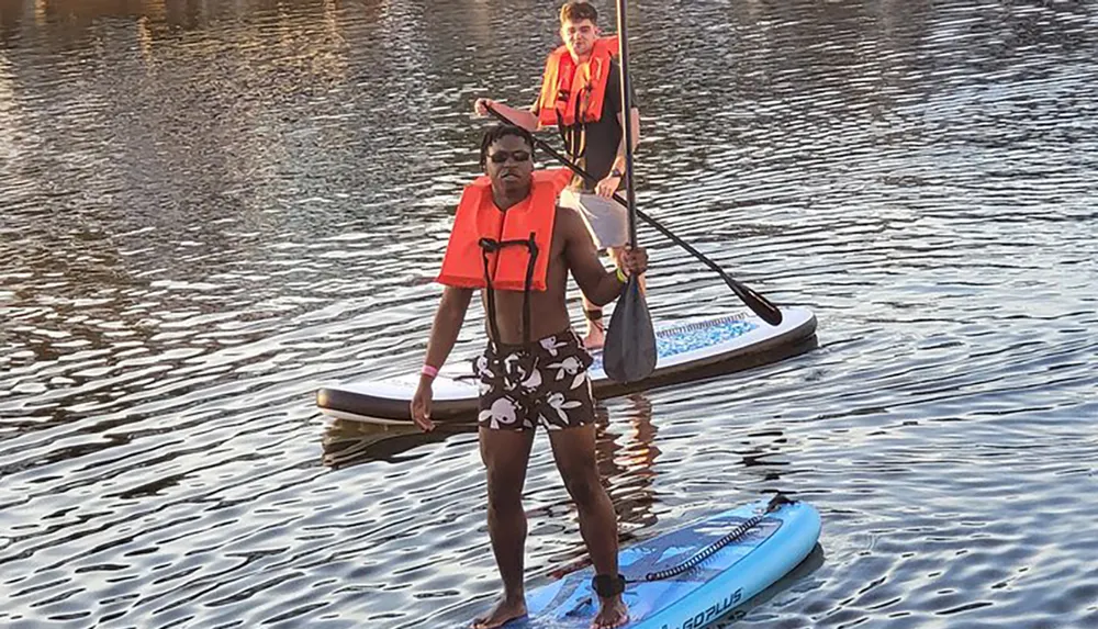 Two individuals are paddleboarding on calm water wearing life jackets and holding paddles during what appears to be late afternoon given the lighting