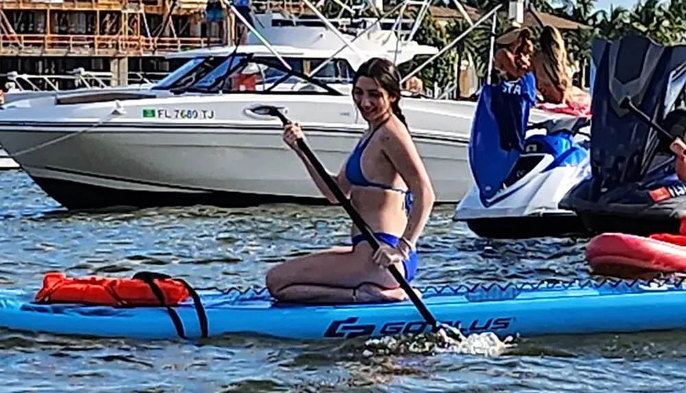 A person is smiling while kneeling on a blue paddleboard holding a paddle with boats and jet skis in the background on a sunny day