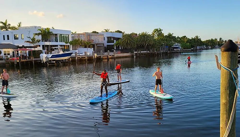 Several people are paddleboarding on a calm waterway with residential buildings and docks in the background during the late afternoon