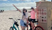 Two people are posing with raised arms next to their bicycles on Hollywood Beach, near a sign listing beach regulations.