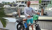 A man in casual clothing is smiling while standing with his hand on the handlebar of a fat tire electric bicycle at a marina with boats docked in the background.