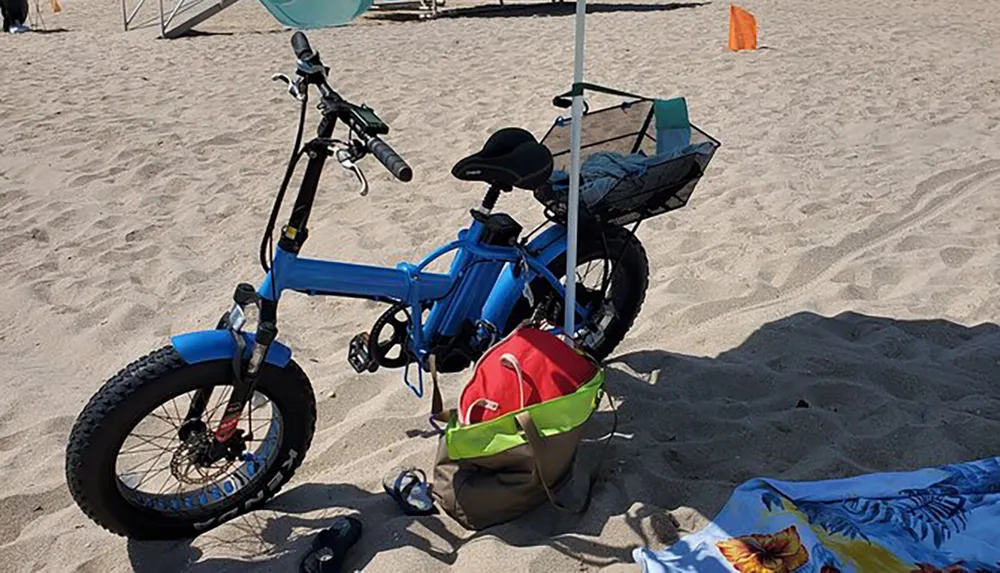 A blue fat-tire electric bike is parked on sandy beach ground with a red and green bag hanging on its handlebars a detached bicycle seat in the foreground and a beach towel lying nearby