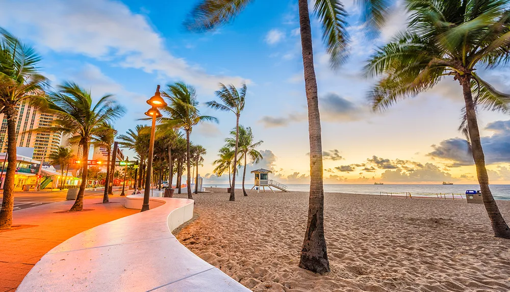 This is a vibrant sunrise scene of a palm-fringed beach promenade with a lifeguard tower overlooking the ocean with ships on the horizon