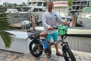 A smiling man stands next to a black electric bicycle with a basket and a green bag on the dock of a marina, with boats in the background.
