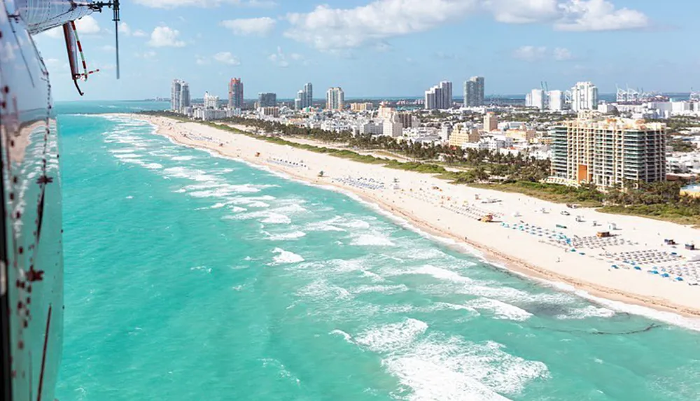 An aerial shot of a sun-drenched Miami beachfront lined with skyscrapers umbrellas and sunbathers viewed from the side of a helicopter