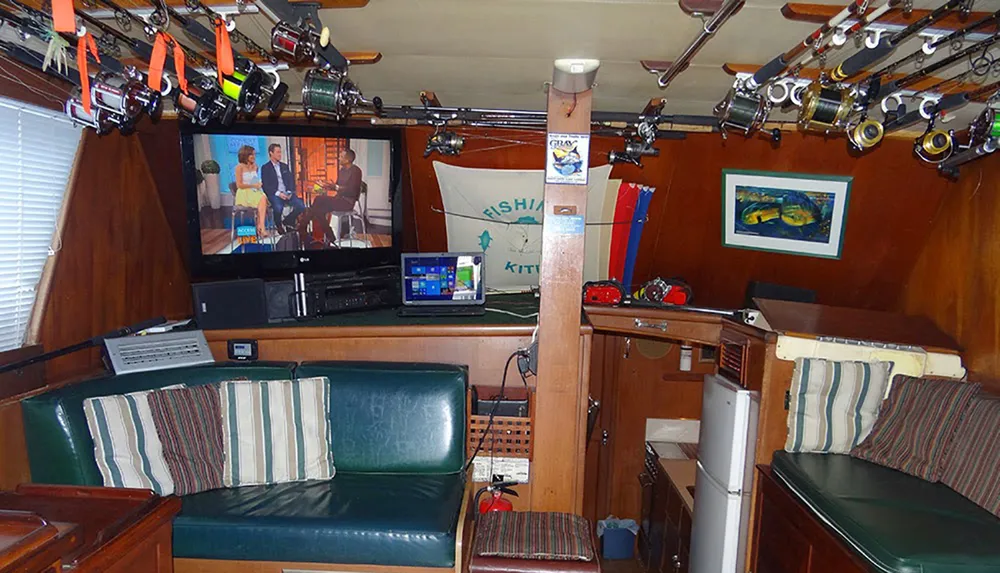 The image shows a cozy interior space with wood paneling lined with numerous fishing rods featuring comfortable seating areas and a TV displaying a talk show