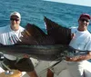 Two smiling men are holding a large sailfish aboard a boat on the ocean