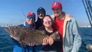 Four people on a boat are smiling with their catch of the day, a large fish, with a backdrop of clear blue skies and distant city buildings.
