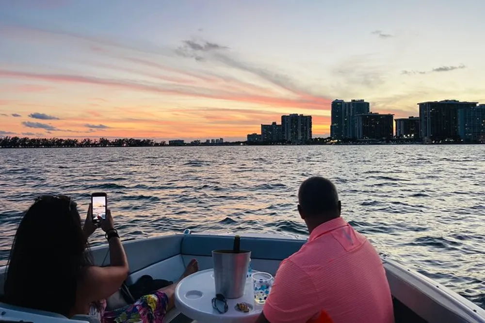 Two people are enjoying a sunset from a boat with one person taking a photo of the scene against the backdrop of a city skyline