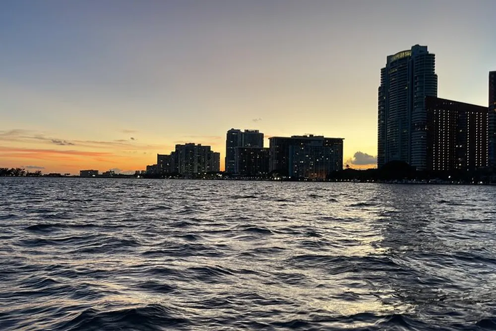 The image shows a city skyline at sunset viewed from across a body of choppy water under a gradient sky transitioning from blue to orange