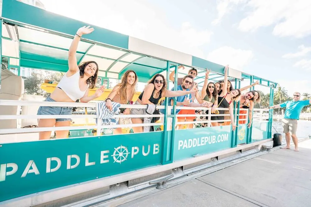 A group of cheerful people are posing and gesturing happily on a bright sunny day aboard a boat with PADDLE PUB written on the side