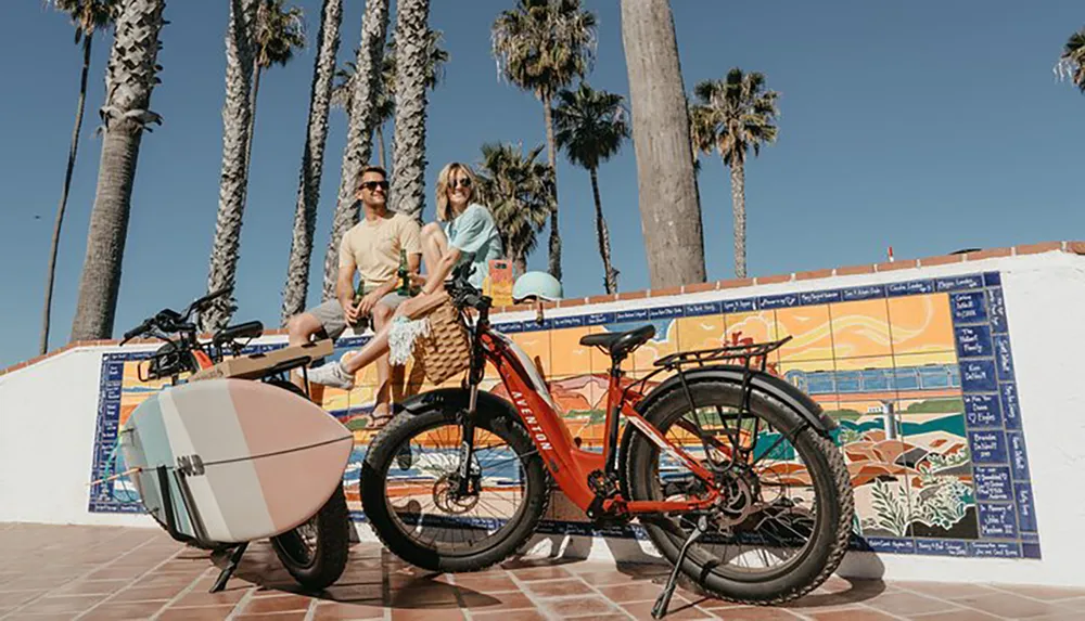 A man and woman relax on a sunlit promenade with palm trees next to a colorful mural an electric bicycle and a surfboard
