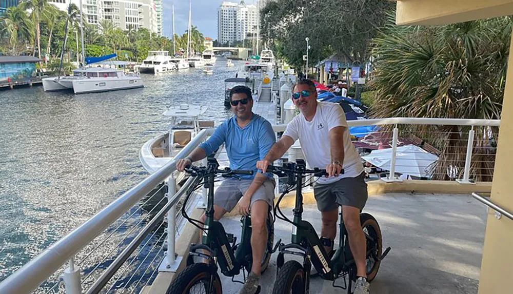 Two people are smiling at the camera while posing with their bicycles on a sunny day by a waterfront filled with boats