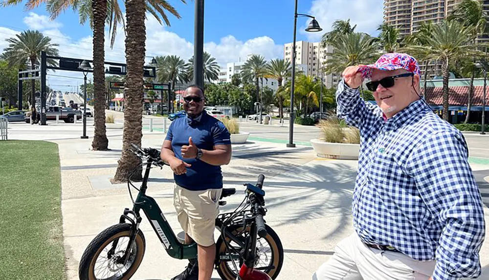Two men are standing on a sunny sidewalk with palm trees in the background one giving a thumbs up next to an electric bicycle and the other shielding his eyes from the sun