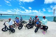 A group of people wearing helmets stands with their fat-tire bicycles on a coastal trail, posing for a photo with a bridge and the ocean in the background.