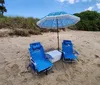 Two blue beach chairs and a cooler are set beneath a patterned umbrella on sandy beach terrain with vegetation in the background