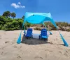 Two blue beach chairs and a cooler are set beneath a patterned umbrella on sandy beach terrain with vegetation in the background