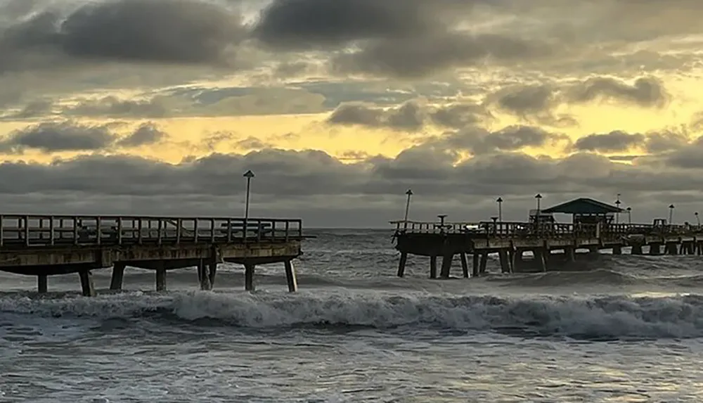 A weathered pier juts into the ocean against a backdrop of dramatic clouds and a setting sun