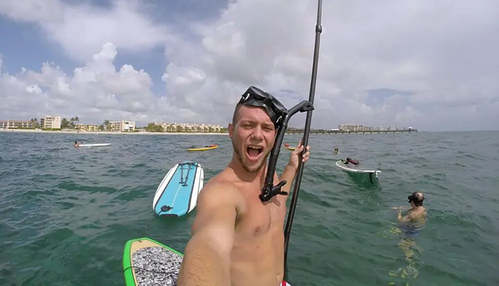 A man takes a selfie with a snorkel mask on his head holding a GoPro on a stick with other people paddleboarding and swimming in the background