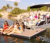 A group of people are enjoying a sunny day on the water with some sitting on a boat and others on a floating mat near the shore