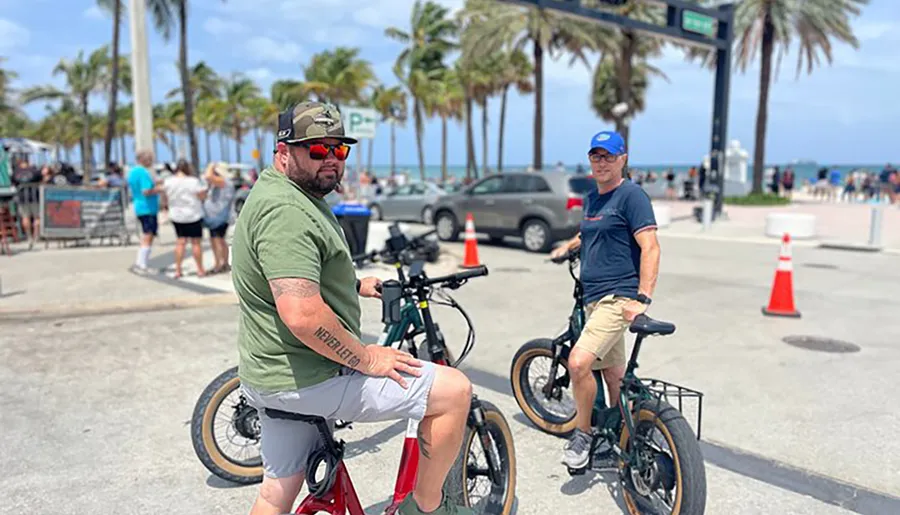 Two men are leisurely riding their bicycles by a beachside promenade lined with palm trees on a sunny day.