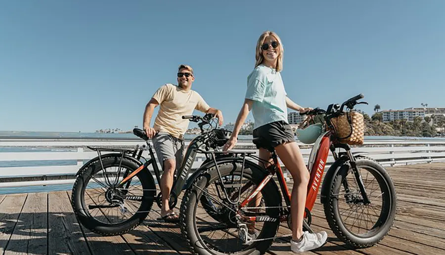 Two people are smiling and standing next to their bicycles on a sunny pier with a clear blue sky and the ocean in the background.